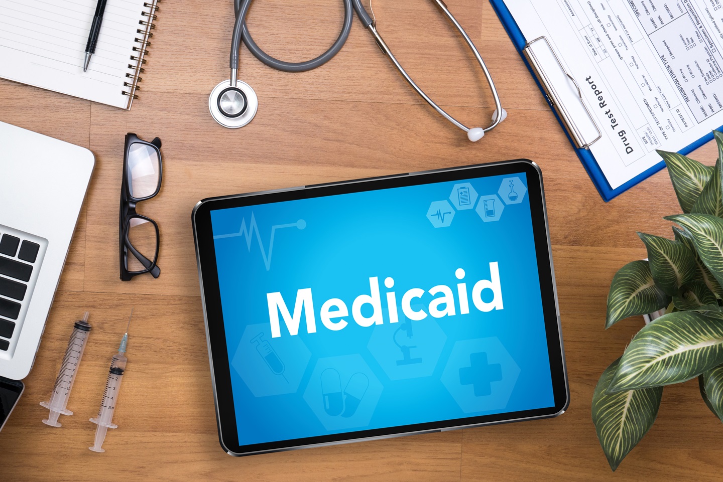 The background is a desk with a notebook, laptop, a pair of glasses, two shots for an injection, in the middle is a tablet screen that says, "Medicaid", and on the top right is a clipboard.